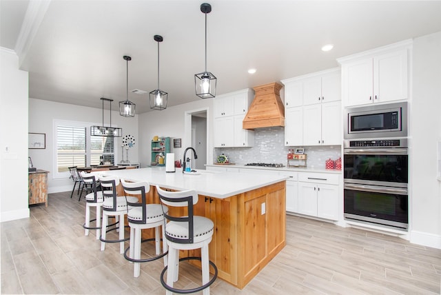 kitchen with premium range hood, hanging light fixtures, a kitchen island with sink, stainless steel appliances, and white cabinets