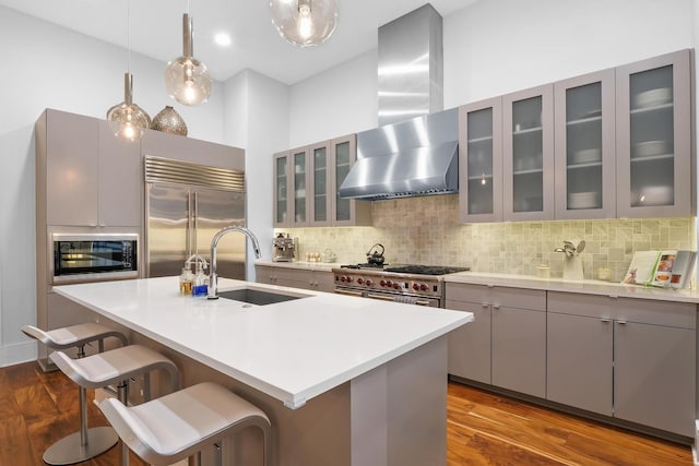 kitchen with wall chimney range hood, built in appliances, decorative backsplash, sink, and a kitchen island with sink