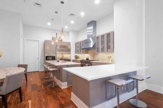 kitchen with decorative light fixtures, wall chimney range hood, a breakfast bar, built in appliances, and an island with sink