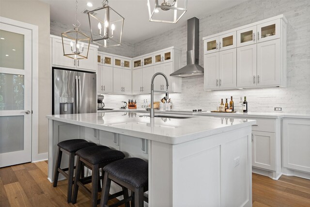 kitchen with stainless steel refrigerator with ice dispenser, wall chimney exhaust hood, an island with sink, and white cabinetry