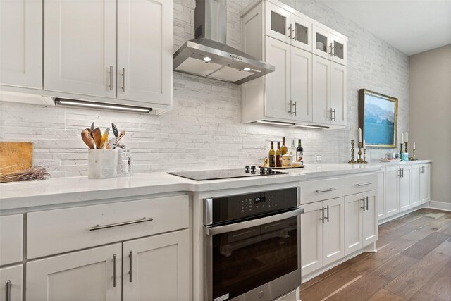 kitchen featuring black electric stovetop, stainless steel oven, white cabinets, and wall chimney range hood