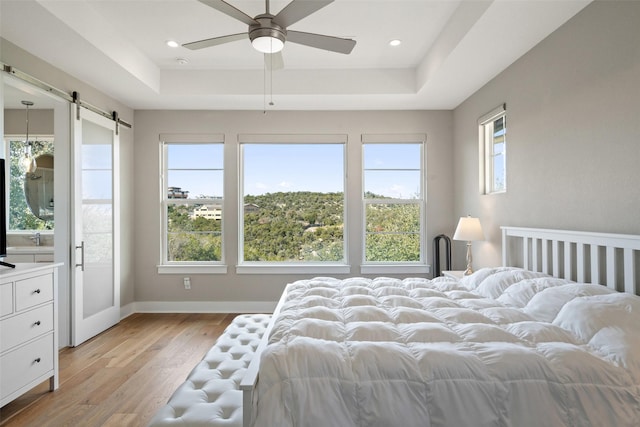 bedroom featuring a barn door, ceiling fan, a raised ceiling, light hardwood / wood-style flooring, and multiple windows