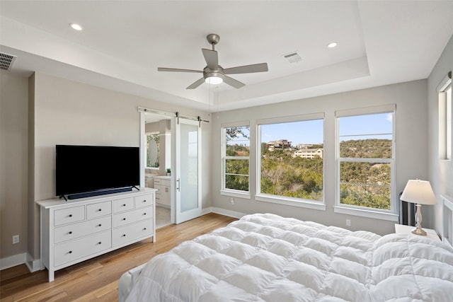 bedroom with light hardwood / wood-style floors, a barn door, ceiling fan, connected bathroom, and a tray ceiling