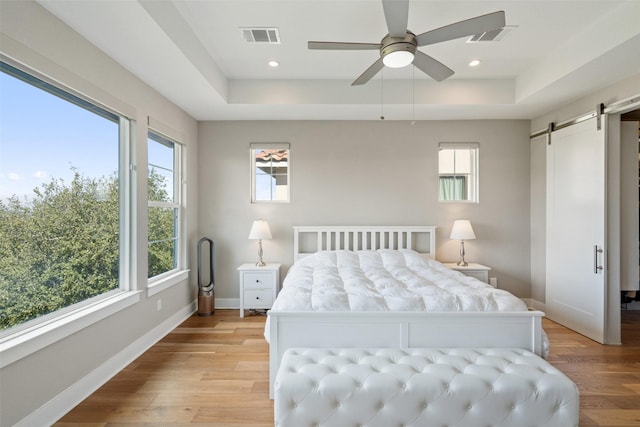 bedroom with ceiling fan, a barn door, a raised ceiling, and light hardwood / wood-style floors