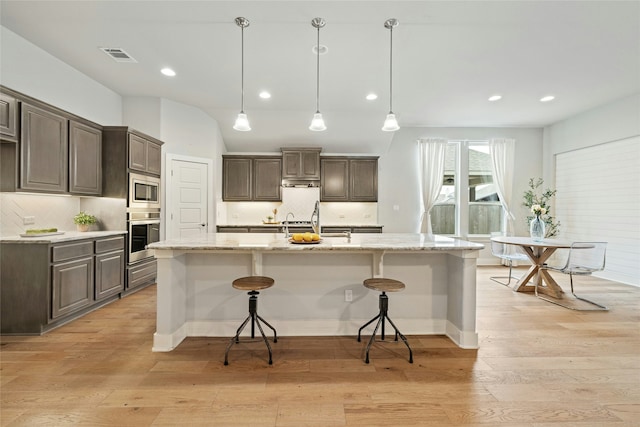 kitchen with appliances with stainless steel finishes, decorative light fixtures, an island with sink, backsplash, and light stone counters