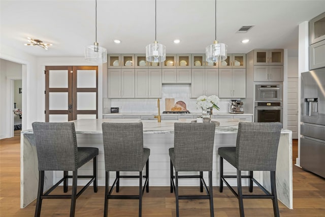 kitchen with tasteful backsplash, decorative light fixtures, an island with sink, stainless steel appliances, and light stone counters