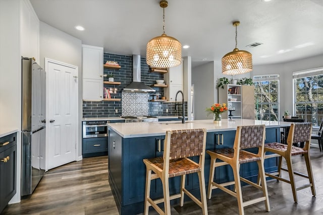 kitchen featuring an island with sink, stainless steel appliances, wall chimney range hood, pendant lighting, and sink