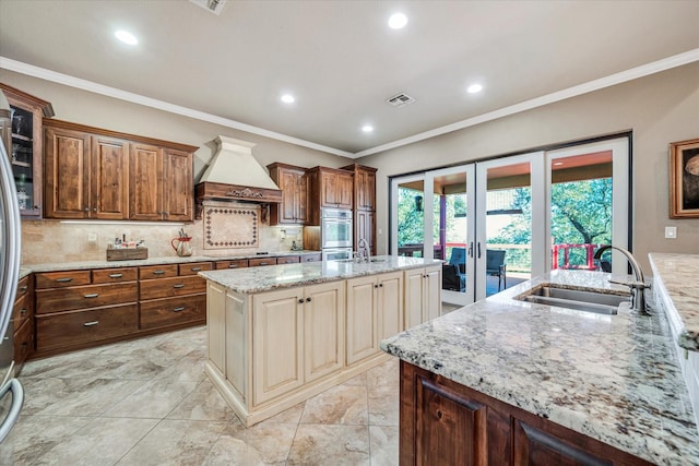 kitchen featuring light stone countertops, custom exhaust hood, french doors, sink, and a kitchen island with sink