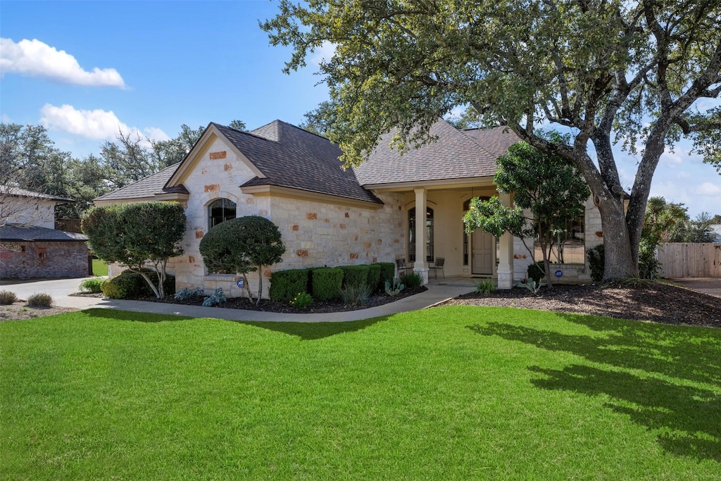 french country inspired facade with stone siding, a front lawn, a shingled roof, and fence