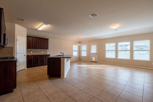 kitchen featuring decorative backsplash, a kitchen island with sink, light tile patterned floors, a notable chandelier, and dark brown cabinetry