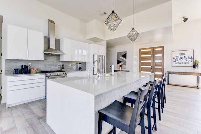 kitchen with white cabinetry, an island with sink, stainless steel appliances, decorative light fixtures, and wall chimney exhaust hood