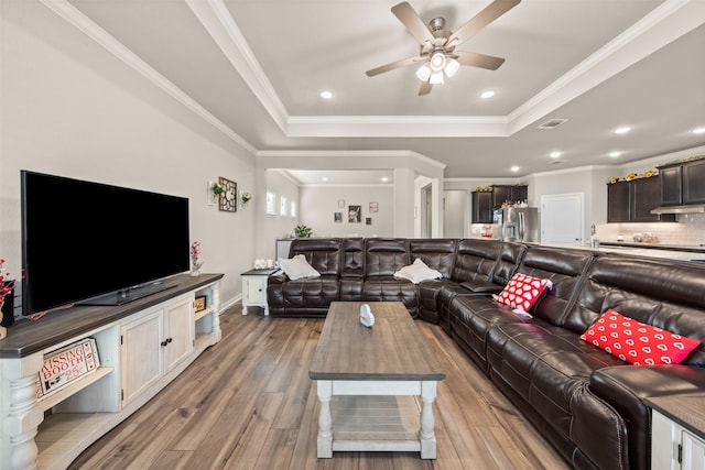 living room featuring a raised ceiling, a ceiling fan, ornamental molding, light wood-style floors, and recessed lighting