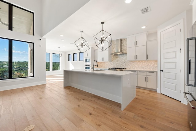 kitchen featuring wall chimney range hood, light hardwood / wood-style floors, white cabinetry, hanging light fixtures, and an island with sink