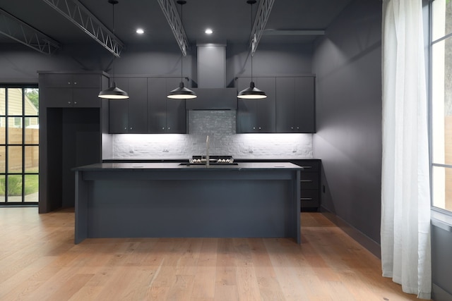 kitchen featuring an island with sink, decorative backsplash, sink, hanging light fixtures, and light hardwood / wood-style flooring