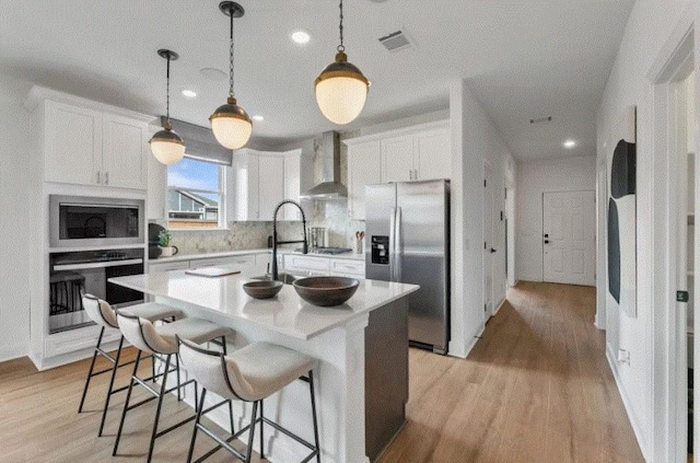 kitchen with stainless steel appliances, decorative light fixtures, a kitchen island with sink, wall chimney exhaust hood, and white cabinets