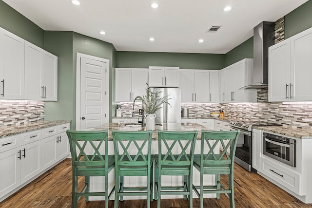 kitchen with wall chimney range hood, white cabinetry, light stone countertops, a breakfast bar area, and stainless steel appliances