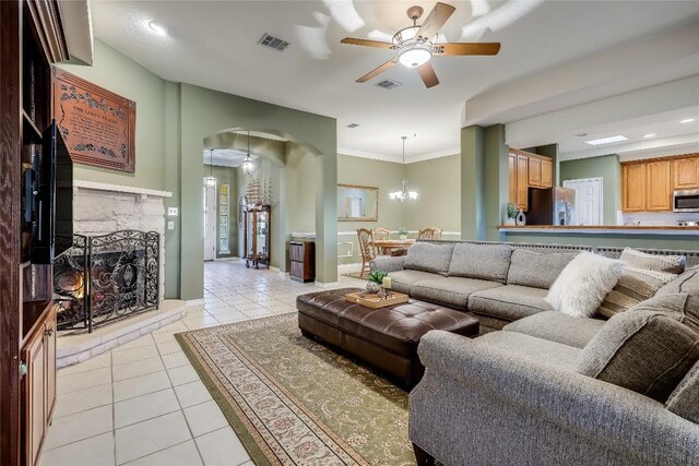 tiled living room with a stone fireplace, ceiling fan with notable chandelier, and crown molding