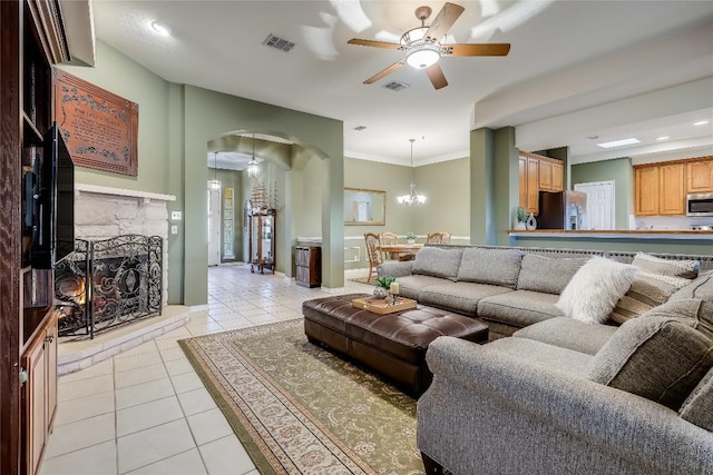 tiled living room featuring crown molding, a stone fireplace, and ceiling fan with notable chandelier