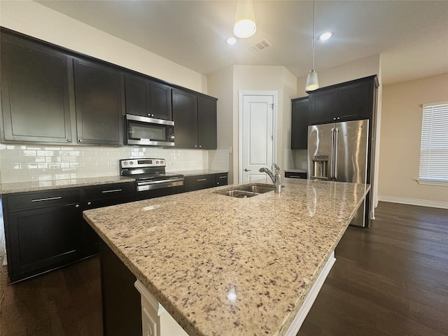 kitchen featuring appliances with stainless steel finishes, a kitchen island with sink, pendant lighting, light stone counters, and sink