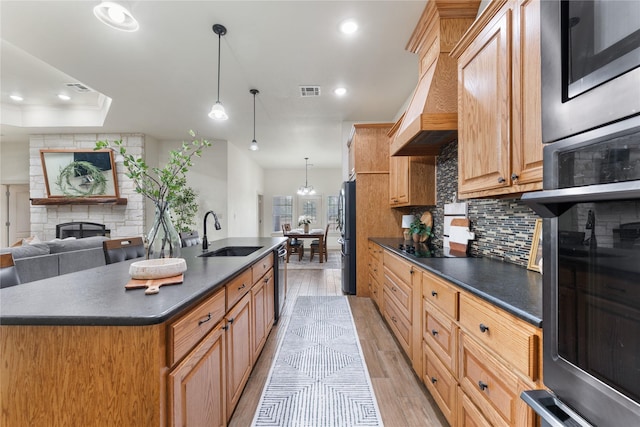 kitchen featuring appliances with stainless steel finishes, a stone fireplace, an island with sink, sink, and light hardwood / wood-style flooring