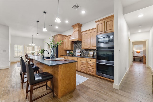 kitchen featuring appliances with stainless steel finishes, decorative light fixtures, a notable chandelier, a center island with sink, and custom range hood