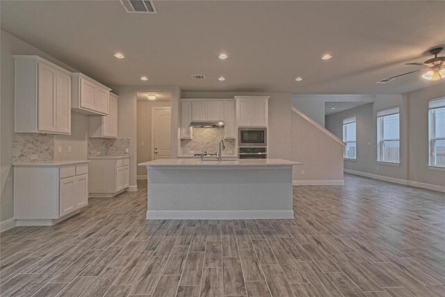 kitchen featuring a kitchen island with sink, white cabinets, built in microwave, oven, and light hardwood / wood-style flooring