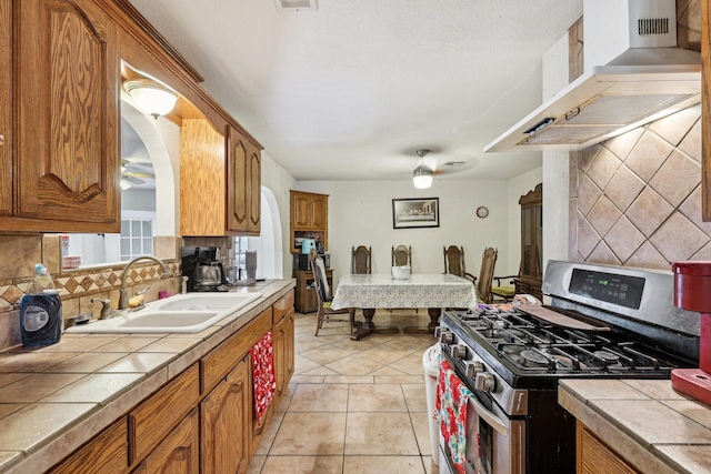 kitchen featuring tasteful backsplash, tile countertops, gas stove, sink, and wall chimney exhaust hood