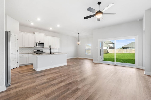 kitchen with white cabinetry, appliances with stainless steel finishes, a kitchen island with sink, light stone countertops, and ceiling fan with notable chandelier