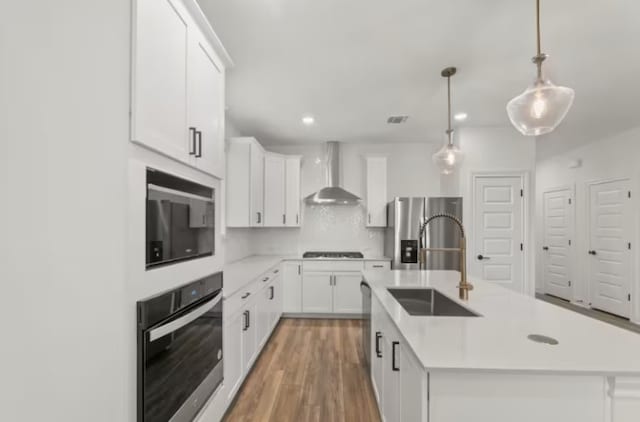 kitchen with wall chimney range hood, a center island with sink, hanging light fixtures, stainless steel appliances, and white cabinets