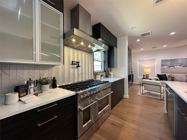 kitchen with light hardwood / wood-style floors, appliances with stainless steel finishes, tasteful backsplash, wall chimney range hood, and light stone counters