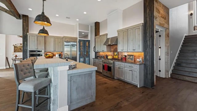 kitchen with a breakfast bar area, a kitchen island with sink, hanging light fixtures, high end appliances, and wall chimney exhaust hood