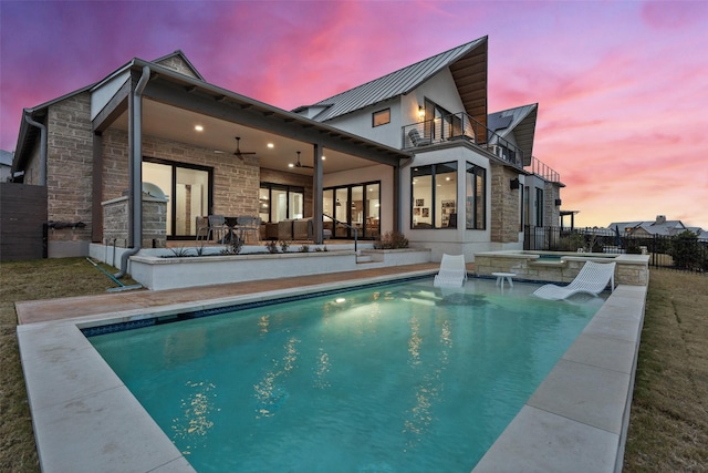 back house at dusk featuring a balcony, a patio area, a swimming pool with hot tub, ceiling fan, and pool water feature