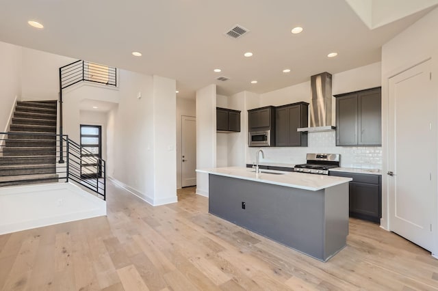 kitchen featuring light hardwood / wood-style flooring, appliances with stainless steel finishes, sink, a kitchen island with sink, and wall chimney range hood