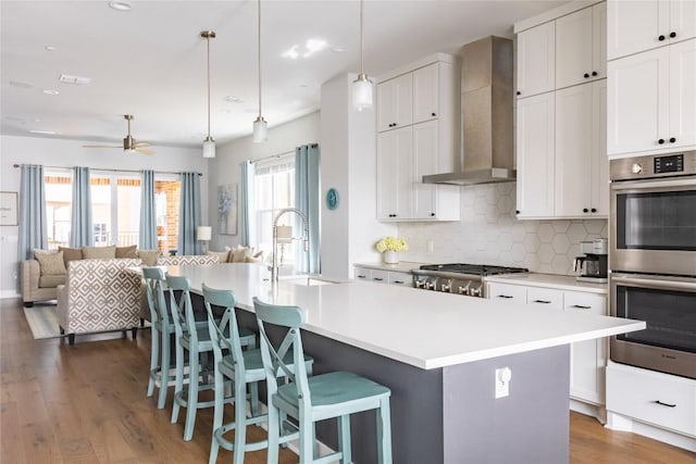 kitchen featuring double oven, wall chimney exhaust hood, a kitchen island with sink, and light countertops