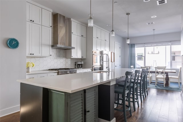 kitchen with pendant lighting, wall chimney range hood, white cabinets, and a large island with sink