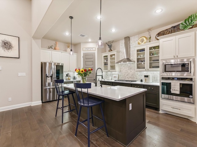 kitchen featuring decorative light fixtures, a center island with sink, wall chimney range hood, stainless steel appliances, and backsplash