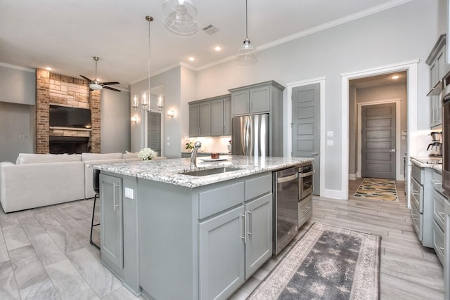 kitchen featuring a stone fireplace, sink, decorative light fixtures, stainless steel appliances, and a center island with sink