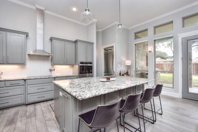 kitchen featuring wall chimney range hood, a center island with sink, oven, and gray cabinetry