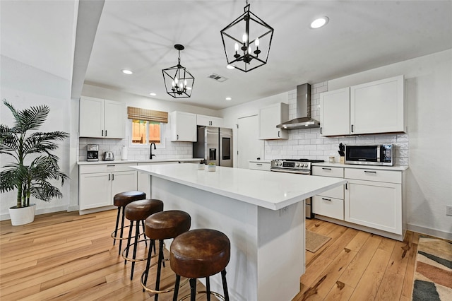 kitchen with wall chimney exhaust hood, white cabinetry, stainless steel appliances, hanging light fixtures, and a center island with sink