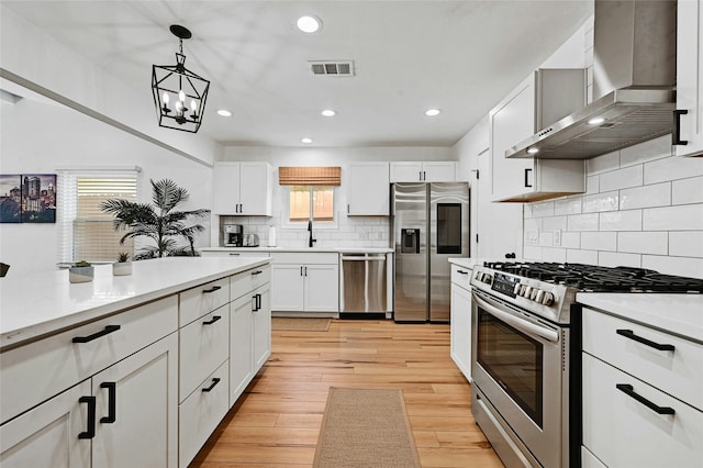 kitchen featuring wall chimney range hood, stainless steel appliances, white cabinetry, and hanging light fixtures