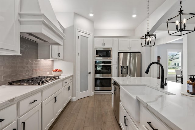 kitchen featuring appliances with stainless steel finishes, custom range hood, white cabinets, light wood-type flooring, and pendant lighting