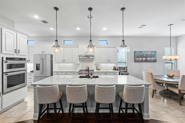 kitchen featuring appliances with stainless steel finishes, white cabinetry, a kitchen island with sink, pendant lighting, and light stone counters