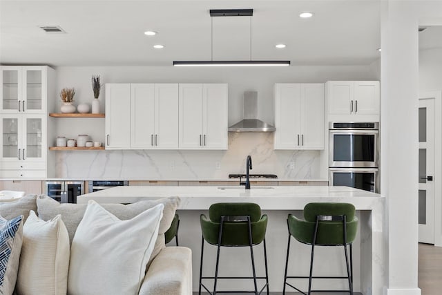 kitchen with stainless steel double oven, white cabinets, a breakfast bar area, and wall chimney exhaust hood