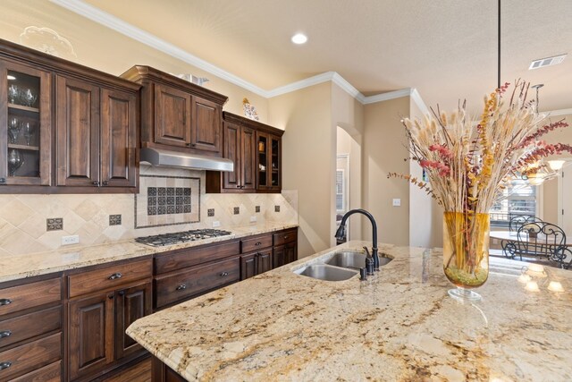 kitchen with glass insert cabinets, under cabinet range hood, stainless steel gas cooktop, a sink, and decorative light fixtures