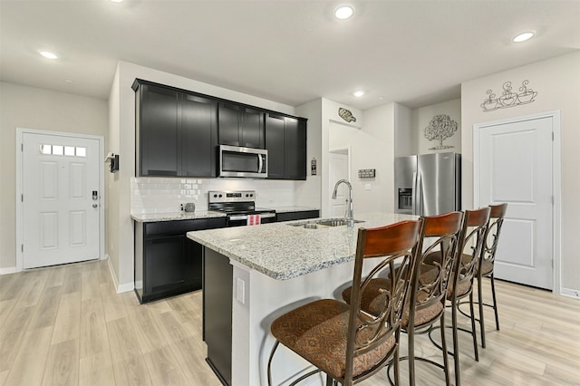 kitchen featuring light stone counters, stainless steel appliances, a kitchen island with sink, a sink, and dark cabinetry