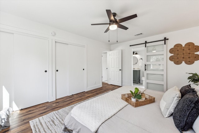 bedroom featuring ensuite bath, dark wood-type flooring, two closets, ceiling fan, and a barn door