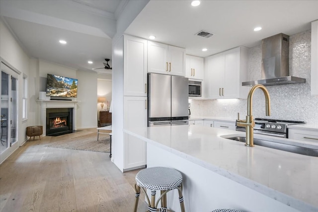 kitchen with stainless steel appliances, a fireplace with flush hearth, visible vents, white cabinets, and wall chimney range hood