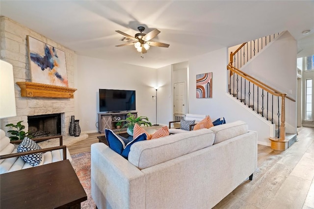 living area with ceiling fan, a stone fireplace, baseboards, stairway, and light wood-type flooring