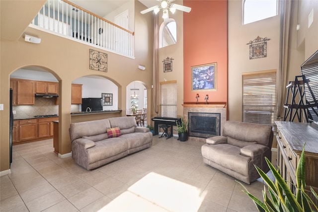 living room featuring a tiled fireplace, ceiling fan, light tile patterned floors, and a towering ceiling