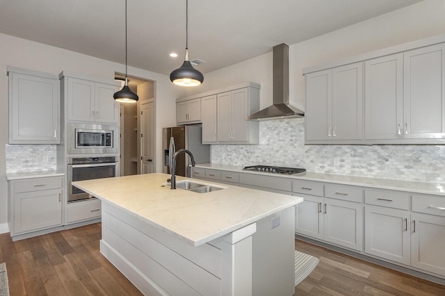 kitchen with wall chimney range hood, stainless steel appliances, sink, and a center island with sink